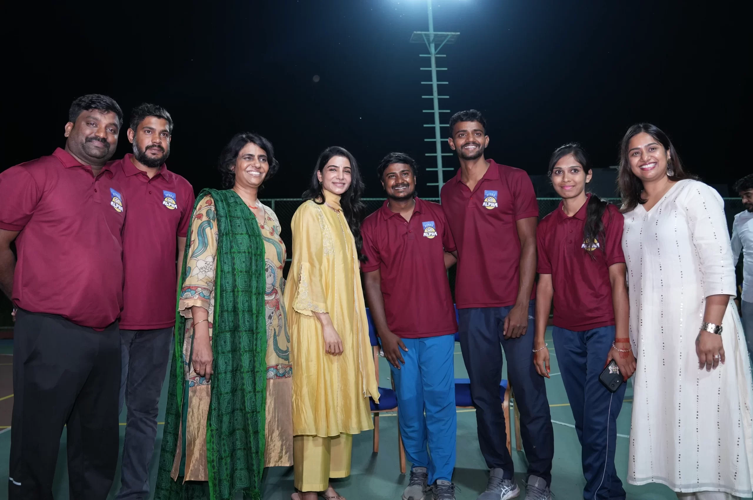 South Indian actress Samantha Ruth Prabhu posing with a group of sports staff in maroon uniforms on an outdoor court at night.